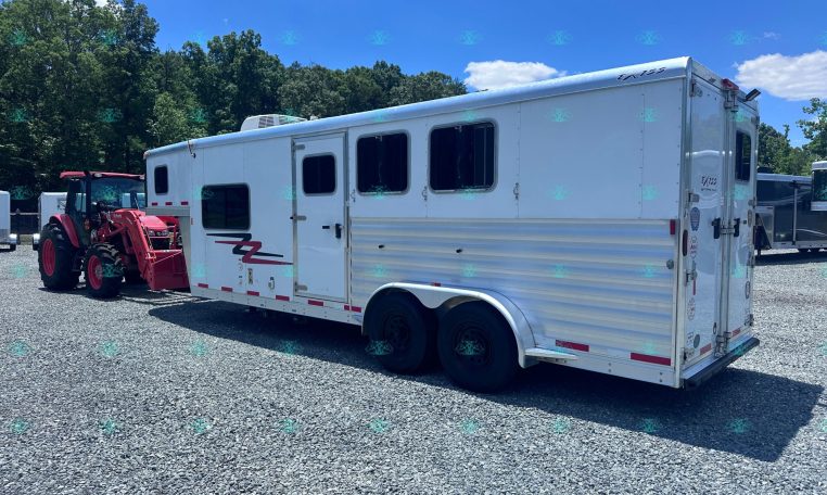 White horse trailer with multiple windows parked on a gravel lot beside a red tractor, against trees and a blue sky.
