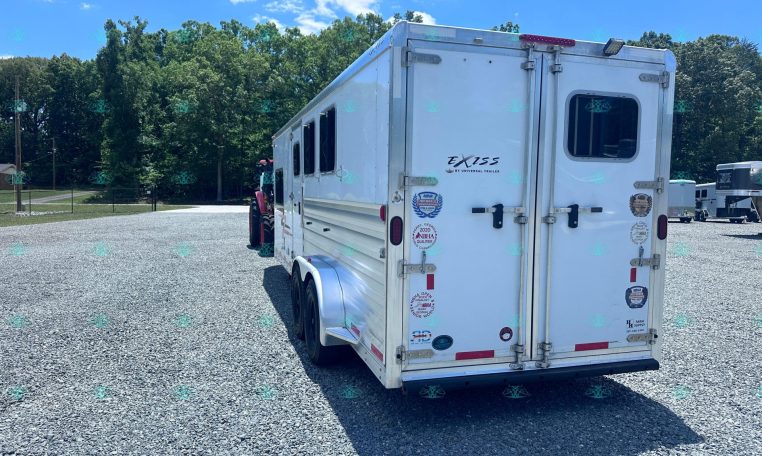 White Exiss horse trailer with dual back doors parked on a gravel lot, trees in the background and a blue sky above