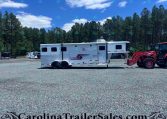 White horse trailer on a gravel lot with a blue sky and tall pine trees in the background; a red tractor attachment is on the right side.