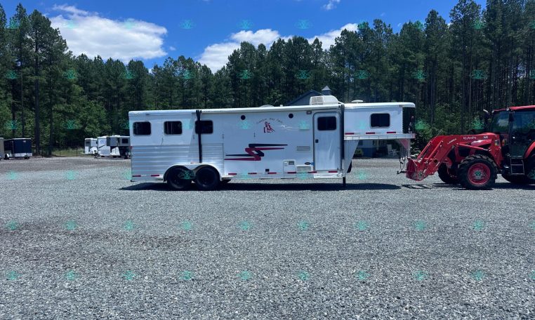 White horse trailer on a gravel lot with a blue sky and tall pine trees in the background; a red tractor attachment is on the right side.
