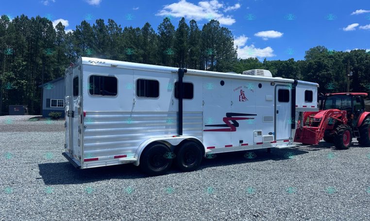 White horse trailer on a gravel lot with a forested backdrop and a red tractor nearby under a bright blue sky.
