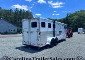 White horse trailer hitched to a red tractor in a gravel lot, with blue sky and trees in the background and a grey building to the left.