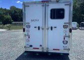 Rear view of a white enclosed trailer parked on a gravel lot, with decals and a small window on the right door.