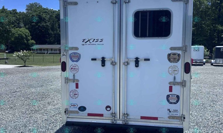 Rear view of a white enclosed trailer parked on a gravel lot, with decals and a small window on the right door.