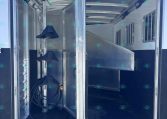 Open aluminum horse trailer interior with divider stall and hose coiled on the floor, bright sky visible outside.