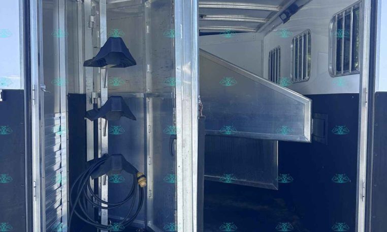 Open aluminum horse trailer interior with divider stall and hose coiled on the floor, bright sky visible outside.