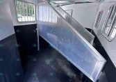 Interior of a silver livestock trailer with a raised metal divider/ramp, aluminum walls, and a barred window on the left side; dark non-slip floor.