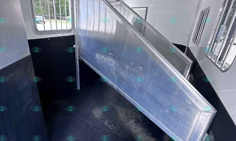 Interior of a silver livestock trailer with a raised metal divider/ramp, aluminum walls, and a barred window on the left side; dark non-slip floor.