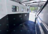 Interior of a aluminum horse trailer with windows along the side and a dark rubber floor, open rear door showing outdoors and other trailers in the background.
