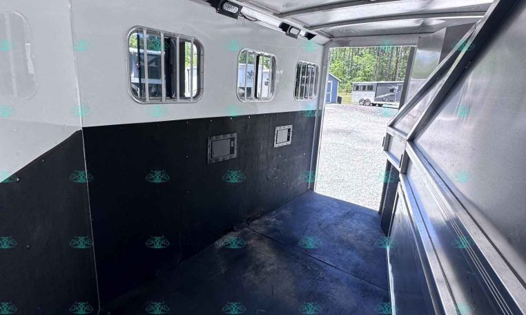 Interior of a aluminum horse trailer with windows along the side and a dark rubber floor, open rear door showing outdoors and other trailers in the background.