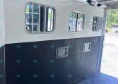 Interior of a horse trailer with white walls, barred windows, and a black lower panel down the side surface.