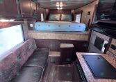 Interior of a compact trailer or RV with a bed at the far end, dark wood cabinets along the left, and a kitchen area on the right side.