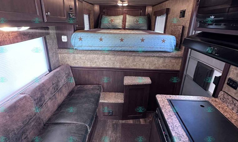 Interior of a compact trailer or RV with a bed at the far end, dark wood cabinets along the left, and a kitchen area on the right side.