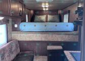 Bedroom in a dark-wood RV with a raised bed, light blue quilted bedding with star accents, and two matching pillows; ceiling HVAC unit and lights above.
