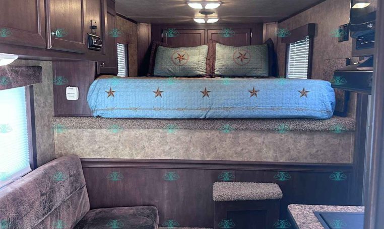 Bedroom in a dark-wood RV with a raised bed, light blue quilted bedding with star accents, and two matching pillows; ceiling HVAC unit and lights above.