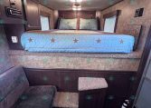Interior RV bedroom with a raised bed, light blue quilt featuring star patterns, two pillows, and dark wood cabinets along the walls.