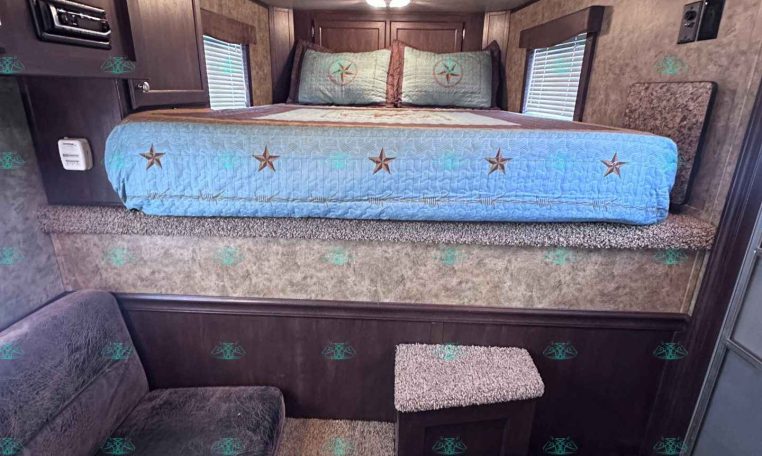 Interior RV bedroom with a raised bed, light blue quilt featuring star patterns, two pillows, and dark wood cabinets along the walls.