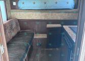 Interior of a compact travel trailer: raised bedroom with a blue quilt, sofa to the left, and a small kitchen/stove area on the right with brown cabinets.