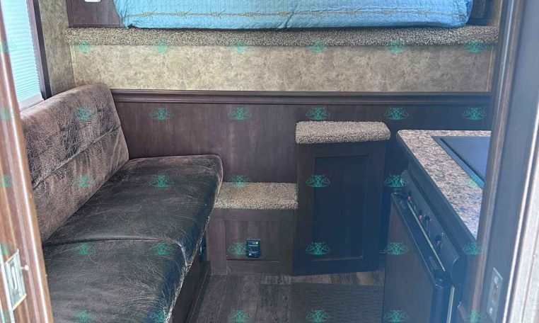 Interior of a compact travel trailer: raised bedroom with a blue quilt, sofa to the left, and a small kitchen/stove area on the right with brown cabinets.