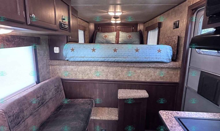 Interior of a trailer showing a raised bed at the back, blue star-pattern bedding, and dark wood cabinets on the sides.
