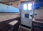 Interior of a compact trailer with a bunk bed over a pine-wood cabinet and stairs beside a small kitchenette. An exterior door and window are visible.