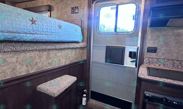 Interior of a compact trailer with a bunk bed over a pine-wood cabinet and stairs beside a small kitchenette. An exterior door and window are visible.