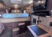 Interior of a compact recreational vehicle with a bed at the rear, blue star-patterned bedding, and warm wood cabinetry along the sides. A kitchen counter with a built-in sink and microwave is on the right, and a seating area with a sofa is in the foreground.