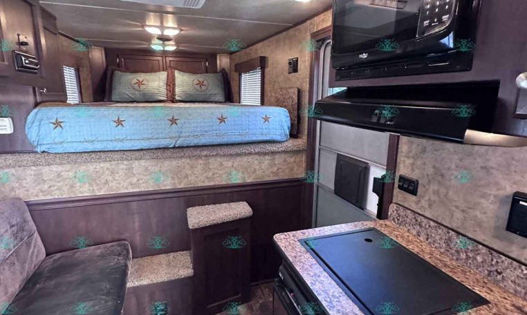 Interior of a compact recreational vehicle with a bed at the rear, blue star-patterned bedding, and warm wood cabinetry along the sides. A kitchen counter with a built-in sink and microwave is on the right, and a seating area with a sofa is in the foreground.
