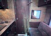 Small kitchen area to the left with sink and counter, a dark wooden door in the center, and a window on the right inside a trailer interior.”,