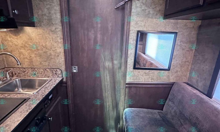 Small kitchen area to the left with sink and counter, a dark wooden door in the center, and a window on the right inside a trailer interior.”,