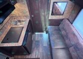 Camper kitchen area with a granite countertop, stainless steel sink, and built-in stove on the left; dark cabinets above. A sofa sits to the right near a window.