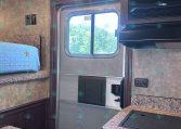 Interior of a small RV/kitchen area with a bunk bed on the left, blue quilt, window in the center, and a microwave above to the right.
