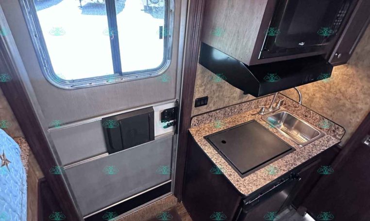 Compact RV kitchen with sink, microwave, and granite countertop beside an exterior exit door windowed by a sliding pane