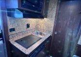 Compact RV kitchen with granite counter, sink, and built-in microwave above a stovetop area in dark wood cabinetry.