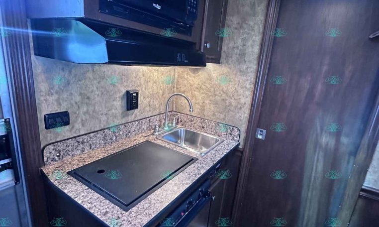 Compact RV kitchen with granite counter, sink, and built-in microwave above a stovetop area in dark wood cabinetry.