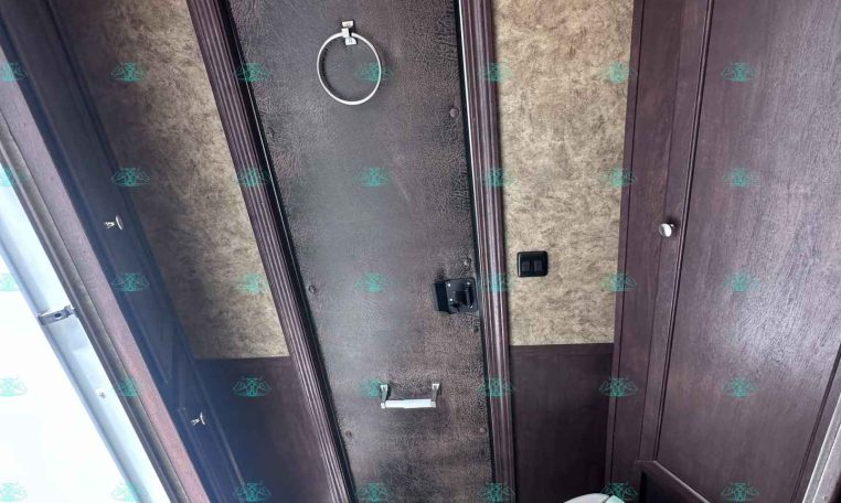 Trailer bathroom with dark wood paneling, beige textured walls, and a frosted door. A circular towel ring hangs on the door above a silver handle.