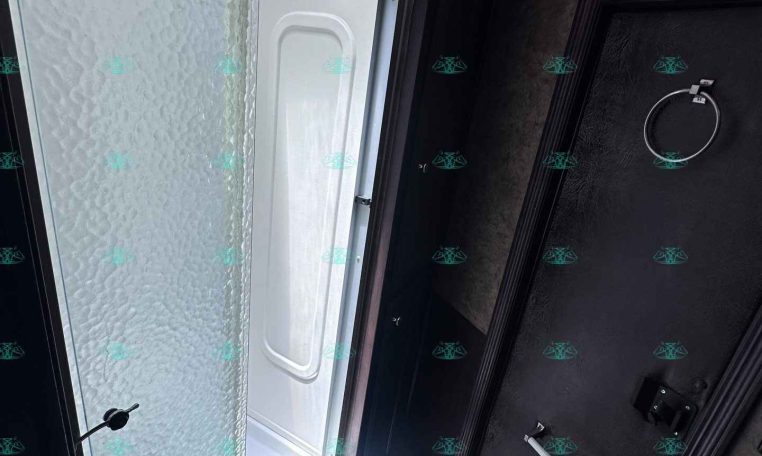 RV bathroom interior featuring a frosted textured glass shower door on the left and a dark wooden door with a towel ring on the right, viewed at a slanted angle.