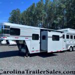 Silver gooseneck horse trailer (Laramie Edition) parked on a gravel lot with tall pine trees in the background, showing multiple windows and storage compartments on the side.
