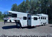 Silver gooseneck horse trailer (Laramie Edition) parked on a gravel lot with tall pine trees in the background, showing multiple windows and storage compartments on the side.