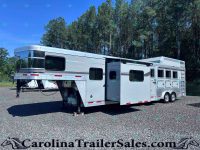 Silver gooseneck horse trailer (Laramie Edition) parked on a gravel lot with tall pine trees in the background, showing multiple windows and storage compartments on the side.