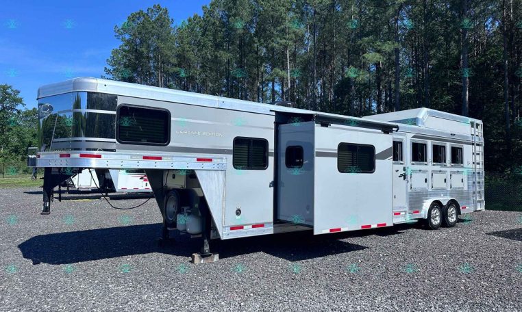 Silver gooseneck horse trailer (Laramie Edition) parked on a gravel lot with tall pine trees in the background, showing multiple windows and storage compartments on the side.