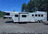 Silver Laramie Edition RV trailer with multiple windows, parked on a gravel lot with a pine forest in the background.