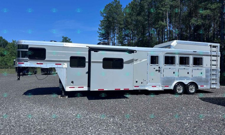 Silver Laramie Edition RV trailer with multiple windows, parked on a gravel lot with a pine forest in the background.