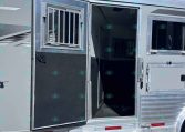 Open doorway of a silver aluminum horse trailer with a barred window, set on a gravel lot with trees in the background