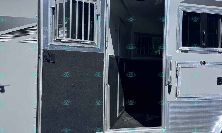 Open doorway of a silver aluminum horse trailer with a barred window, set on a gravel lot with trees in the background