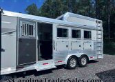 Silver aluminum horse trailer with an open side door, multiple windows, and a metal ladder, parked on gravel with trees in the background