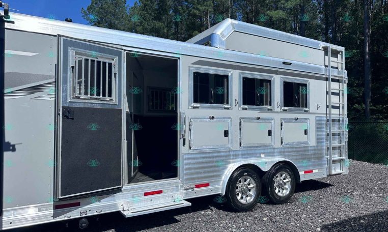 Silver aluminum horse trailer with an open side door, multiple windows, and a metal ladder, parked on gravel with trees in the background