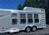 Silver multi-window horse trailer with barred windows, aluminum trim, ladder on the side, parked on gravel with trees and blue sky in background.