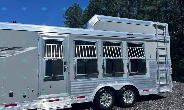 Silver multi-window horse trailer with barred windows, aluminum trim, ladder on the side, parked on gravel with trees and blue sky in background.