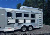 Silver aluminum horse trailer with three side windows, a rear ladder, and chrome trim parked on gravel with trees in the background.